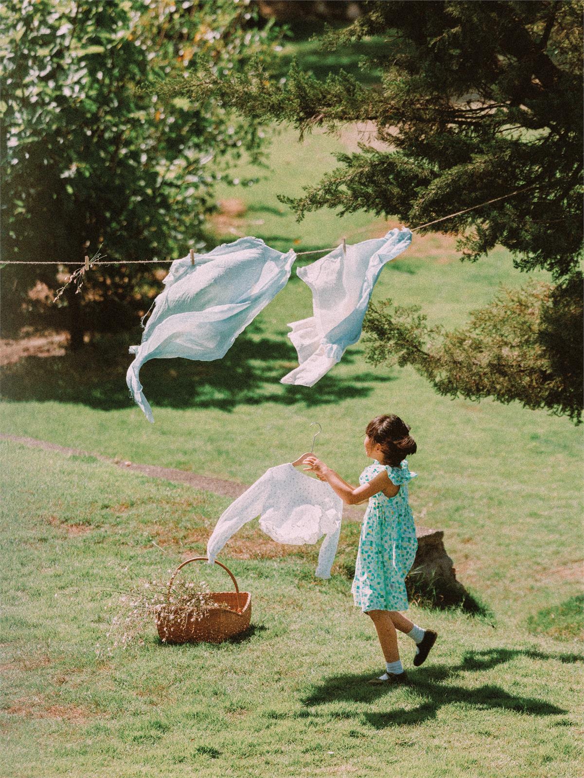 A child carrying white laundry across a sunlit garden