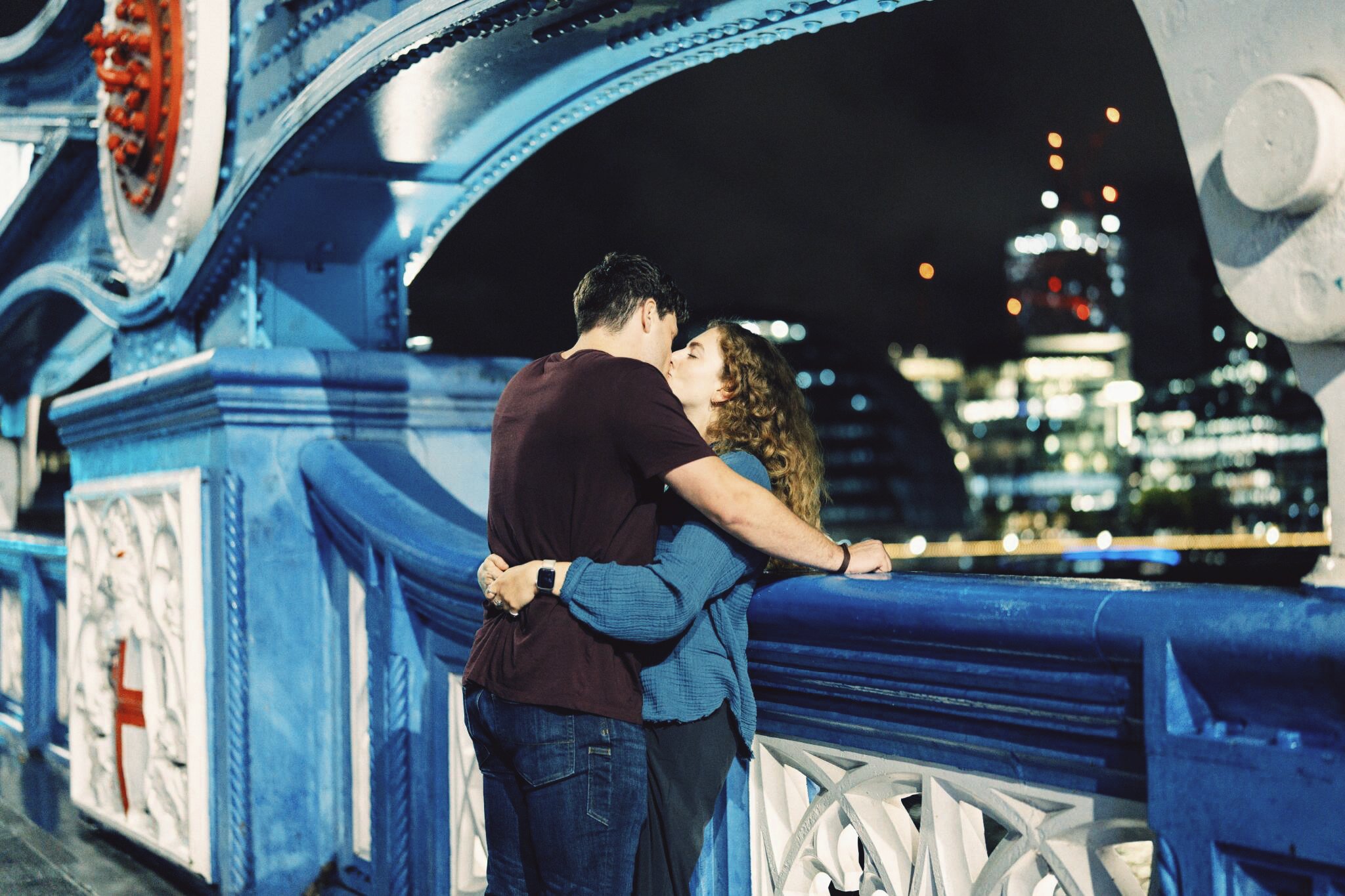 A couple embracing on a blue bridge at night, city lights behind