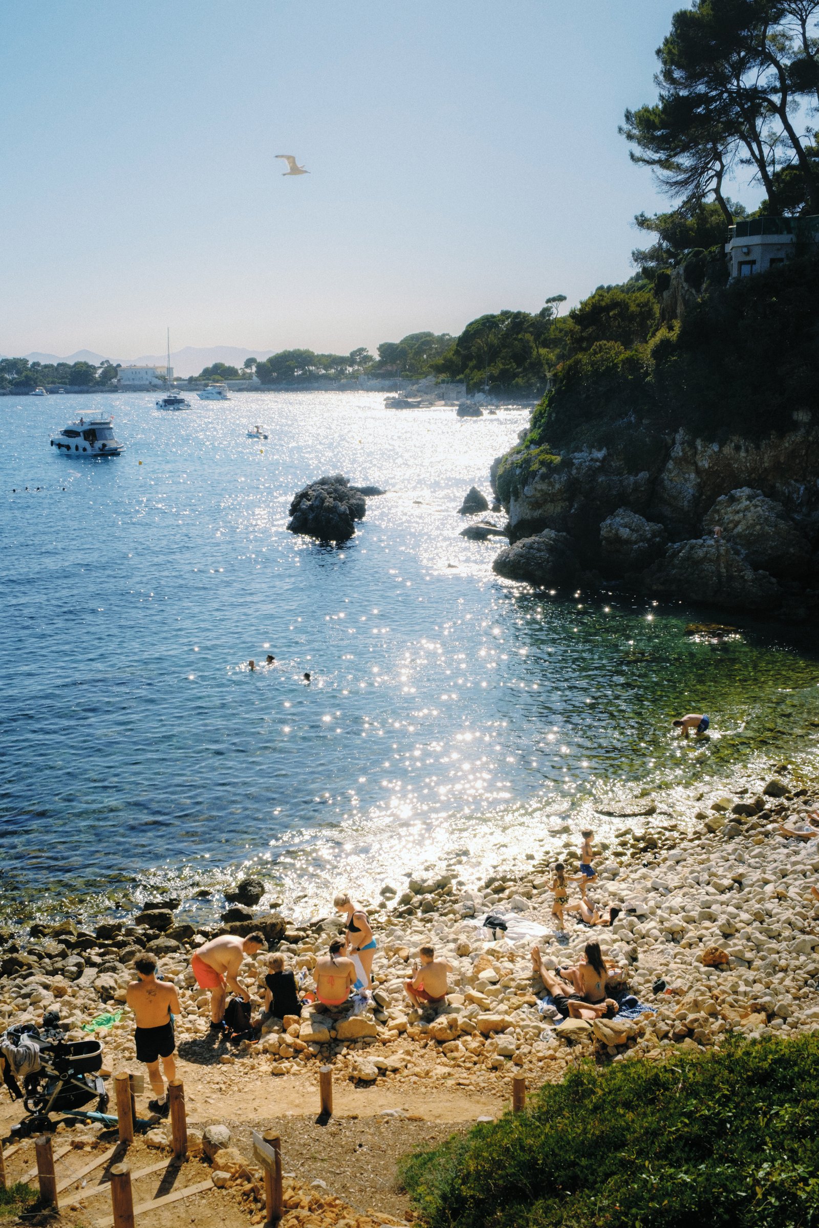 Sunbathers on a rocky beach cove with boats in bright sunlight